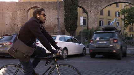Young hipster man riding bike in Rome city centre with historic ruins and cars wearing sunglasses on sunny day slow motion camera car steadycam