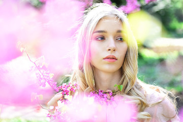 Fototapeta premium Spring bloom concept. Young woman enjoy flowers in garden, defocused, close up. Girl on dreamy face, tender blonde near violet flowers of judas tree, nature background. Lady in park on spring day.