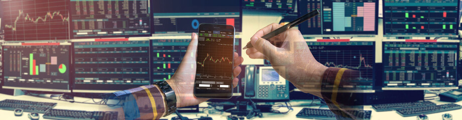 Double exposure of hand holding smartphone with Stock market quotes and chart in monitor computer room with business office equipments .business and money concept, panorama