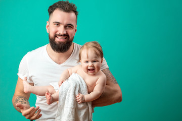 Young happy bearded father in white t-short holding his laughing infant daughter wrapped in towel in arms, studio shot on blue background.