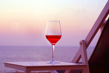 A glass of red wine standing on a table near a summer chair against a background of a sea horizon at sunset. A glass of red wine against the background of the evening sea horizon at sunset.