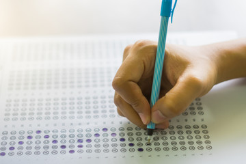 Close up of high school or university student holding a pen writing on answer sheet paper in examination room. College students answering multiple choice questions test in testing room in university.