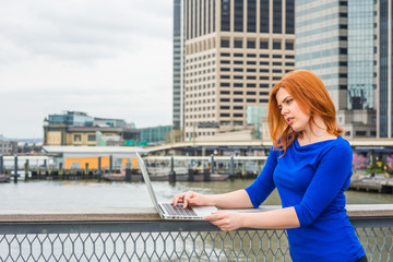 Young American Woman traveling, working in New York, wearing blue long sleeve T shirt, standing by river at harbor in business district of Manhattan, looking down, working on laptop computer..