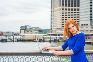 Fototapeta na wymiar Young American Woman traveling, working in New York, wearing blue long sleeve T shirt, standing by river at harbor in business district of Manhattan, working on laptop computer, looking away, thinking