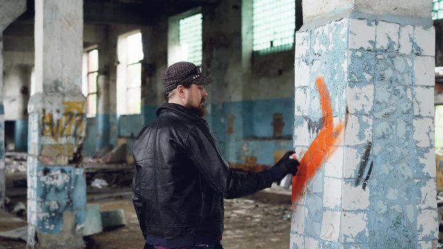 Bearded graffiti artist is painting on pillar in old abandoned building using aerosol paint. Modern youth subculture, creative people and street art concept.