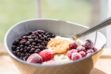 Macro closeup of oatmeal oat grain rice pudding porridge bowl with peanut butter, frozen berries, raspberries, blueberries, strawberries on table by window, spoon