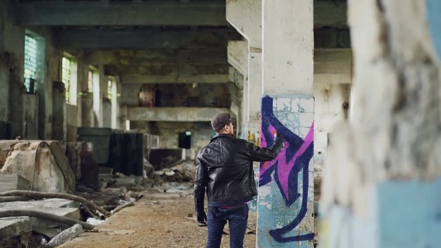 Male graffiti painter is creating abstract image with spray paint inside abandoned empty building. Old column is in foreground, dirty walls and windows in background.