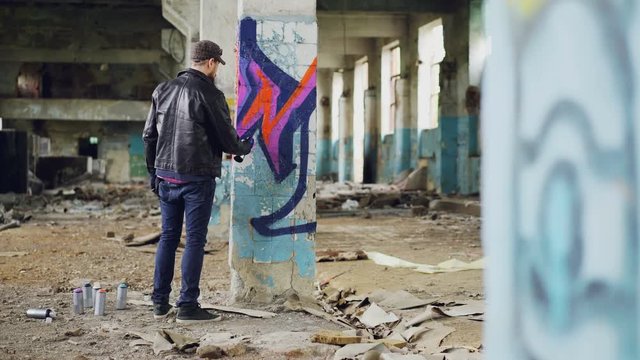Bearded young man is painting graffiti inside old industrial buliding using aerosol paint, he is making bright abstract image on column. Creativity and people concept.