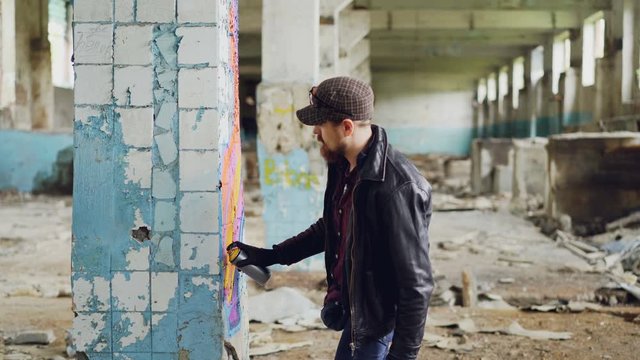 Handsome bearded guy is holding aerosol paint and drawing graffiti on pillar inside spacious abandoned house. Creative people, empty buildings and modern art concept.