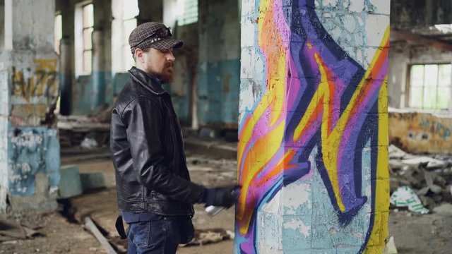 Professional graffiti painter is creating abstract image on large pillar inside abandoned building with spray paint. Young man is wearing leather jacket, cap and gloves.