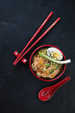 Red tableware with chicken ramen noodles, vertical shot on a black stone background, view from above with space