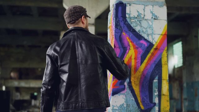 Rear view of male graffiti artist in leather jacket painting on damaged column inside empty industrial building. Young people, creativity, casual clothing and modern art concept.