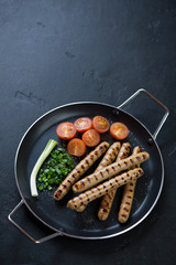 Metal pan with grilled pork sausages, tomatoes and green onion, elevated view on a black stone background, copyspace