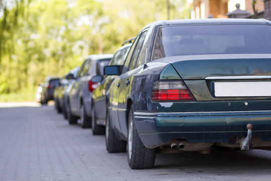 Long row of cars parked in quiet neighborhood on clean empty paved street on background of beautiful green trees on bright sunny summer or spring day.