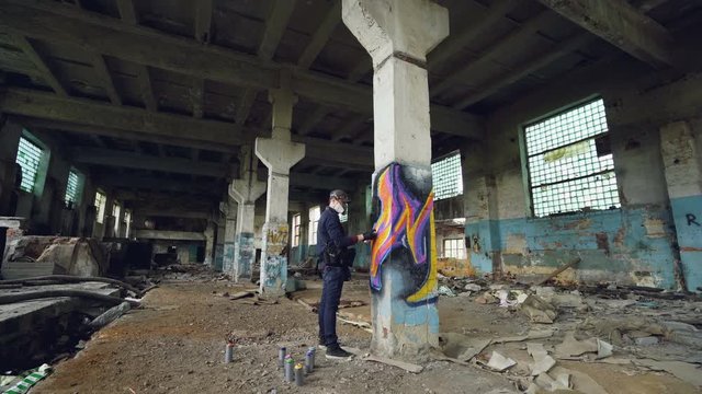 Graffiti artist in protective mask is painting on high column in abandoned industrial building. Creative people, modern wall art and protective equipment concept.