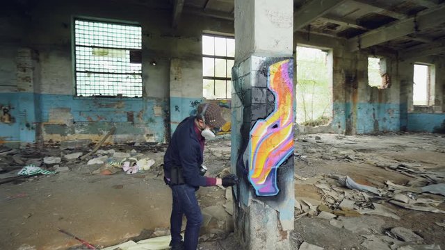 Pan shot of adult man graffiti artist in protective face mask and gloves painting on high pillar inside damaged empty industrial building. Creativity and people concept.