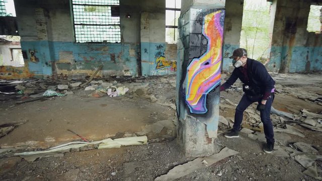 Pan shot of masked graffiti artist drawing abstract images on pillar in large empty building using paint spray. Painter is wearing casual clothing and protective gloves.