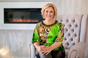 Fototapeta na wymiar Delightful blonde woman in colorful green shirt and pants, sitting in chair on background of fireplace