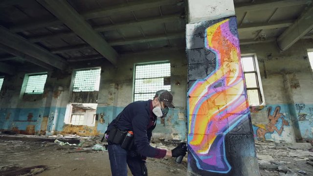 Urban street artist is painting graffiti in abandoned building with dirty walls and windows, he is using paint spray. Modern artwork and creative people concept.