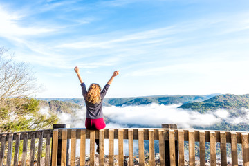 Back of young woman sitting on railing by mountains and floating fog clouds in morning in Grandview...