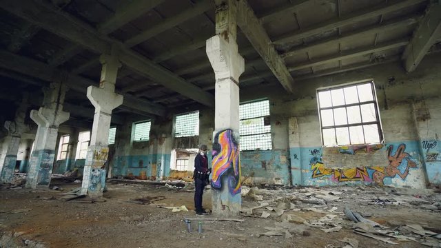 Male graffiti artist in respirator is shaking spray paint then painting on high pillar inside dirty empty building. Beautiful abstract image on column is visible.