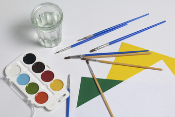 Watercolors and brushes on a white surface. Next glass for water and paper. Subjects for creativity.