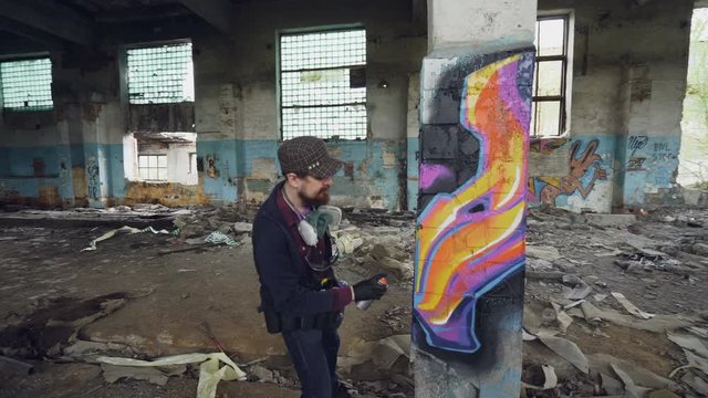 Pan shot of damaged abandoned building with high columns inside and male graffiti artist drawing abstract images on pillar. Man has protective gloves and gas mask.