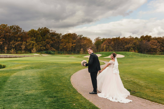 The bride and groom walking on the field with green grass. Autumn forest in the background. beautiful sky with clouds