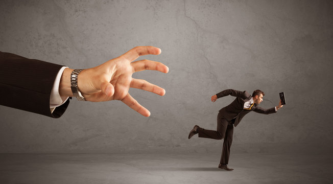 Young miniature businessman running from a big hand with grey background