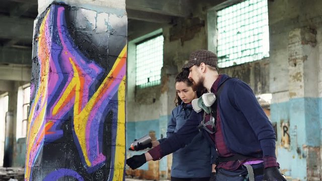Professional graffiti artist is teaching pretty girl to paint with aerosol spray paint standing together inside adandoned warehouse near old column and talking.
