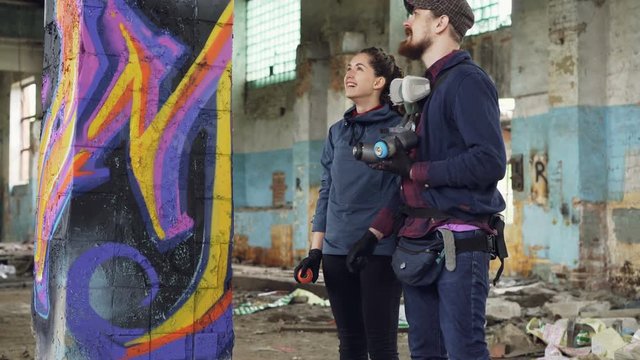 Attractive girl is learning how to create graffiti under guidance of professional artist handsome bearded guy. Young people are talking and laughing painting with spray paint.