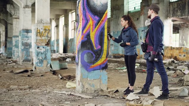 Professional graffiti painter is teaching pretty girl to work with paint spray while working together in adandoned building. Young people are wearing casual clothing.