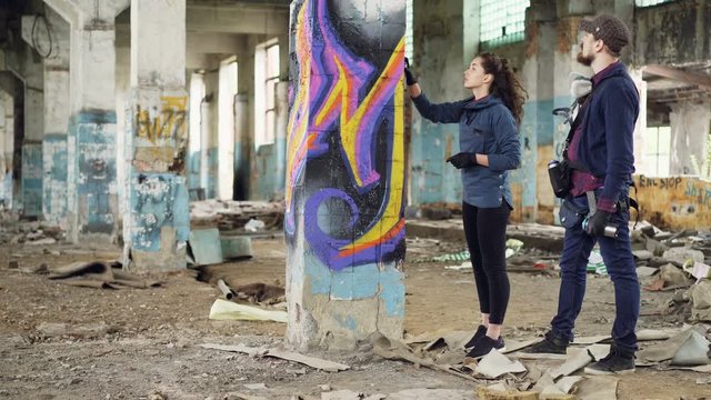 Handsome bearded man graffiti artist is teaching his female friend to draw with spray paint standing inside old damaged building and talking holding aerosol cans.