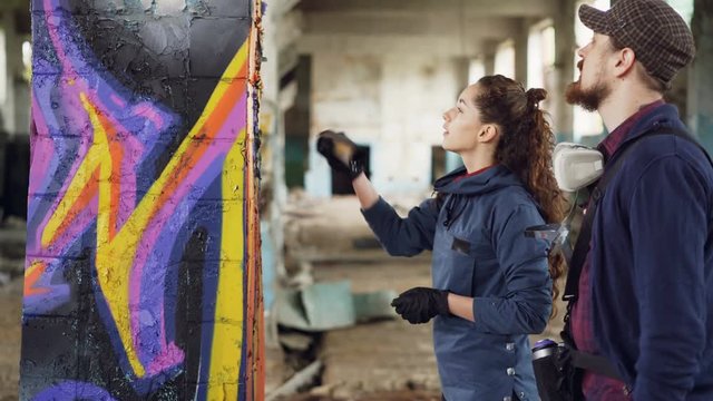 Cheerful young woman amateur graffiti artist is learning to work with spray paint from skilled bearded painter while decorating old column in empty warehouse.