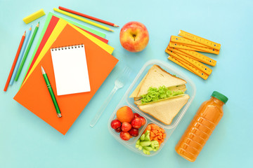 School supplies and lunch box with sandwiches, pieces of cucumbers and carrots, apricot, cherries, apple, bottle of juice on light blue background, back to school concept.