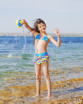 A cool young girl in a swimsuit with long hair flying in the wind watering from a watering can standing in sea water