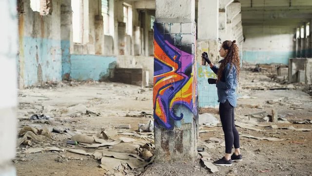 Pretty girl graffiti artist is decorating old damaged column inside empty industrial building with abstract pictures. Modern painter is using aerosol spray paint.