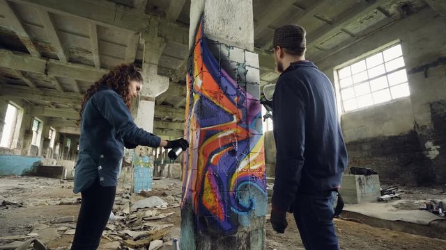 Low angle shot of graffiti painters decorating abandoned industrial building with bright images using aerosol paint. Dirty old walls and high ceiling are visible.