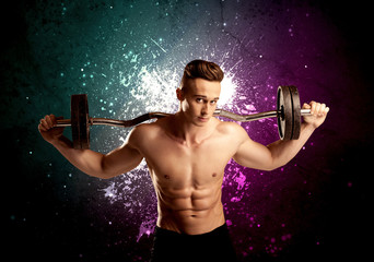 A sexy male fitness trainer showing his muscles and looking seductive with a weight in his hands in front of bright paint splash purple wall concept.