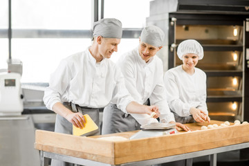 Three young bakers in uniform forming dough for baking on the wooden table standing together at the...