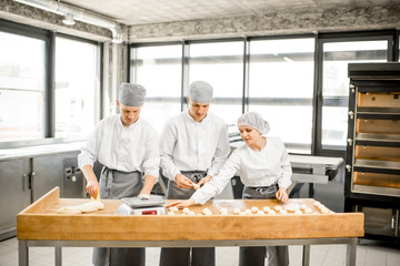 Three young bakers in uniform forming dough for baking on the wooden table standing together at the modern manufacturing