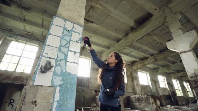 Attractive girl urban artist is painting graffiti in abandoned building with dirty walls and windows, she is using paint spray. Modern artwork and creative people concept.