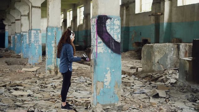 Urban artist is decorating column in abandoned warehouse with abstract image using aerosol paint. Girl is wearing casual clothing and listening to music with headphones.