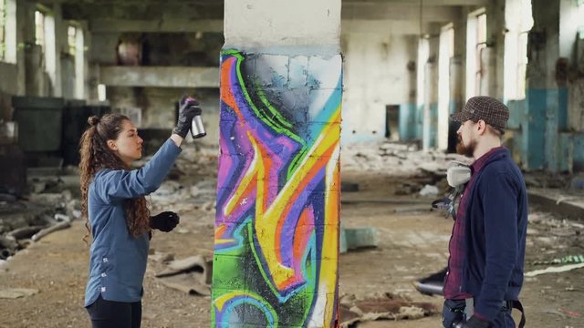 Creative team of two urban painters are drawing graffiti with spray paint while decorating old industrial warehouse with destroyed dirty walls and windows.