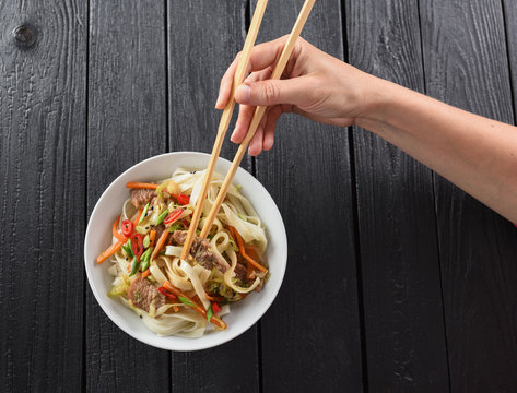 Slender woman hand with chop sticks in bowl of traditional Asian udon noodles on black background minimalist style