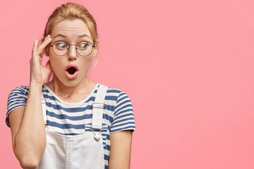 Indoor shot of stunned blonde woman with blue eyes, keeps mouth wide opened, being puzzled by something, wears denim overalls and sailor t shirt, stands against pink background with blank space