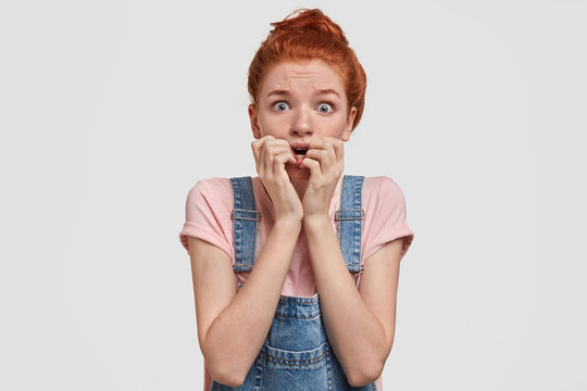 Frightened beautiful freckled female bites finger nails nervously, afraids of speaking in front of audience, feels puzzled, wears jean dungareess, poses against white background. Omg, what is it?