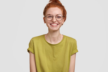 Glad smiling ginger female with white teeth, dressed in casual clothing, wears glasses, enjoys free time with friends, isolated over white background. Foxy delighted teenager being in good mood