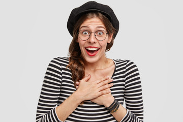Emotive beautiful young French woman looks with stunned expression, being in good mood, wears striped jacket with beret, feels surpirised by something pleasant, poses against white background