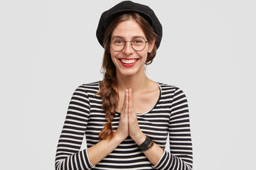 Happy young Parisian female with glad expression, has broad smile, makes praying gesture, asks friend to go for walk during nice weather, wears beret and stiped jacket, isolated on white background